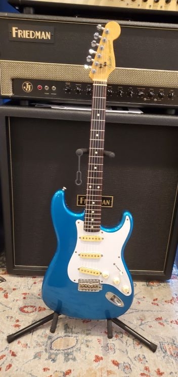 blue strat with white pickguard on stand in front of amp