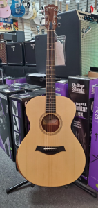 Taylor Academy 12E acoustic guitar sitting on a guitar stand in front of purple and black boxes